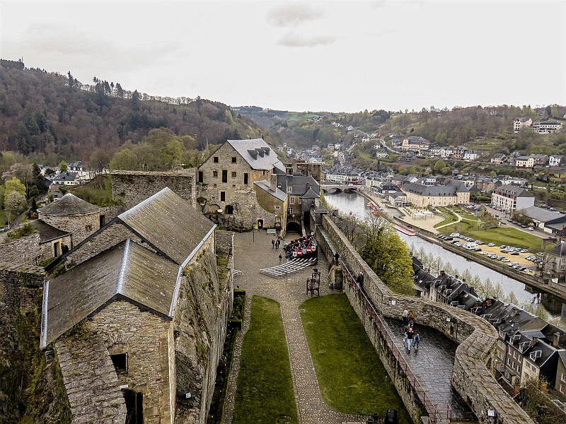 
Aerial view of an old stone castle complex with surrounding walls, a courtyard, and buildings, set on a hillside overlooking a river, trees, and a town with houses and streets. The Bouillon Castle in Belgium - Wallonia