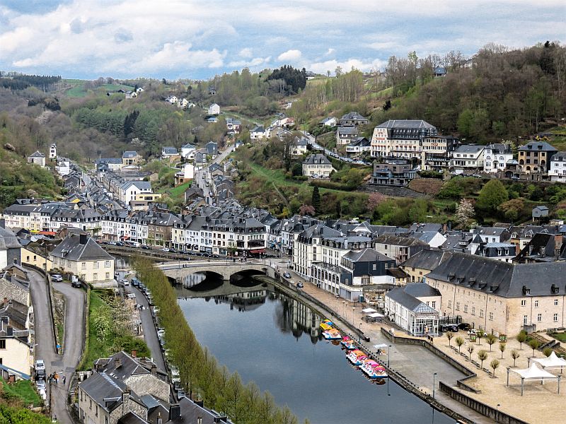 A small European town with clusters of white and gray buildings, situated along a curving river with boats docked by the shore, a stone bridge, hilly greenery, and forested hills in the background. View from the Bouillon castle and the River Semois, Belgium, Wallonia