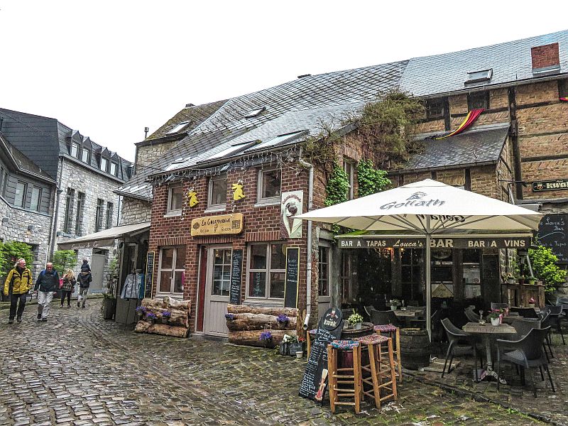 A cobblestone street with people walking by a rustic brick building housing a bar and tapas restaurant; outdoor seating is set up under a large white umbrella, and the building is decorated with plants and signs. Durbuy, Belgium, Wallonia