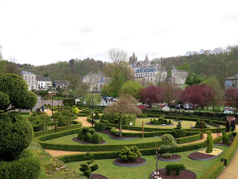  
A formal garden with neatly trimmed shrubs and hedges, winding gravel paths, and topiary designs is set in front of a stone building and castle on a hill, with trees and parked cars in the background. Topiary park in Durbuy, Belgium, Wallonia