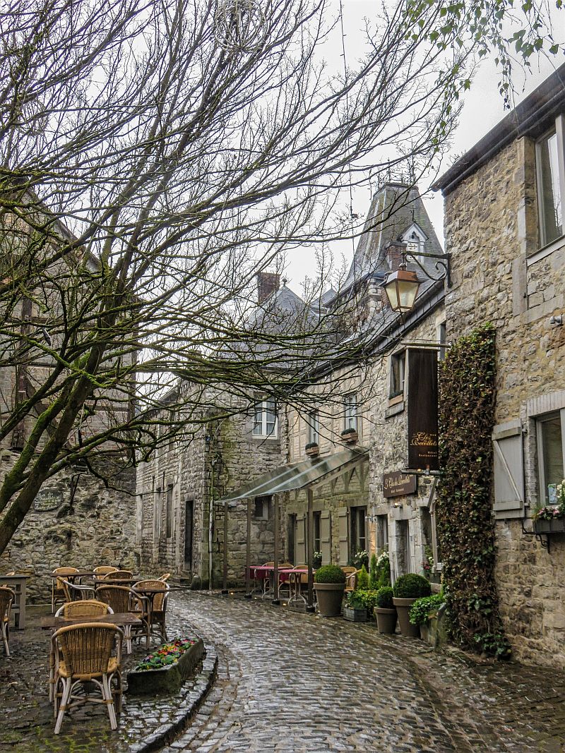 A narrow cobblestone street lined with stone buildings and outdoor café tables on a cloudy day; a leafless tree stretches over the path, and potted plants decorate the storefronts. Durbuy, Belgium