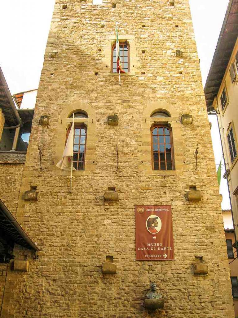 A rectangular stone building with arched windows and flags. A large red sign reads "Museo Casa di Dante" above a small sculpted bust and plaque at the base of the wall. Casa di Dante Museum in Florence