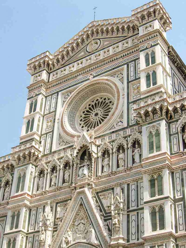 The image shows the ornate marble facade of Florence Cathedral, featuring intricate carvings, a large rose window, pointed arches, and statues set in niches against a clear blue sky.