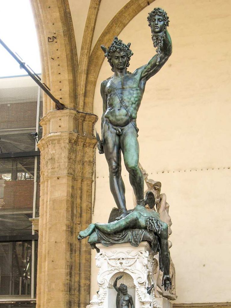A bronze statue of Perseus holding Medusa’s severed head in his left hand and a sword in his right, standing over Medusa’s body, displayed outdoors under an arched structure. Perseus of Cellini in Florence
