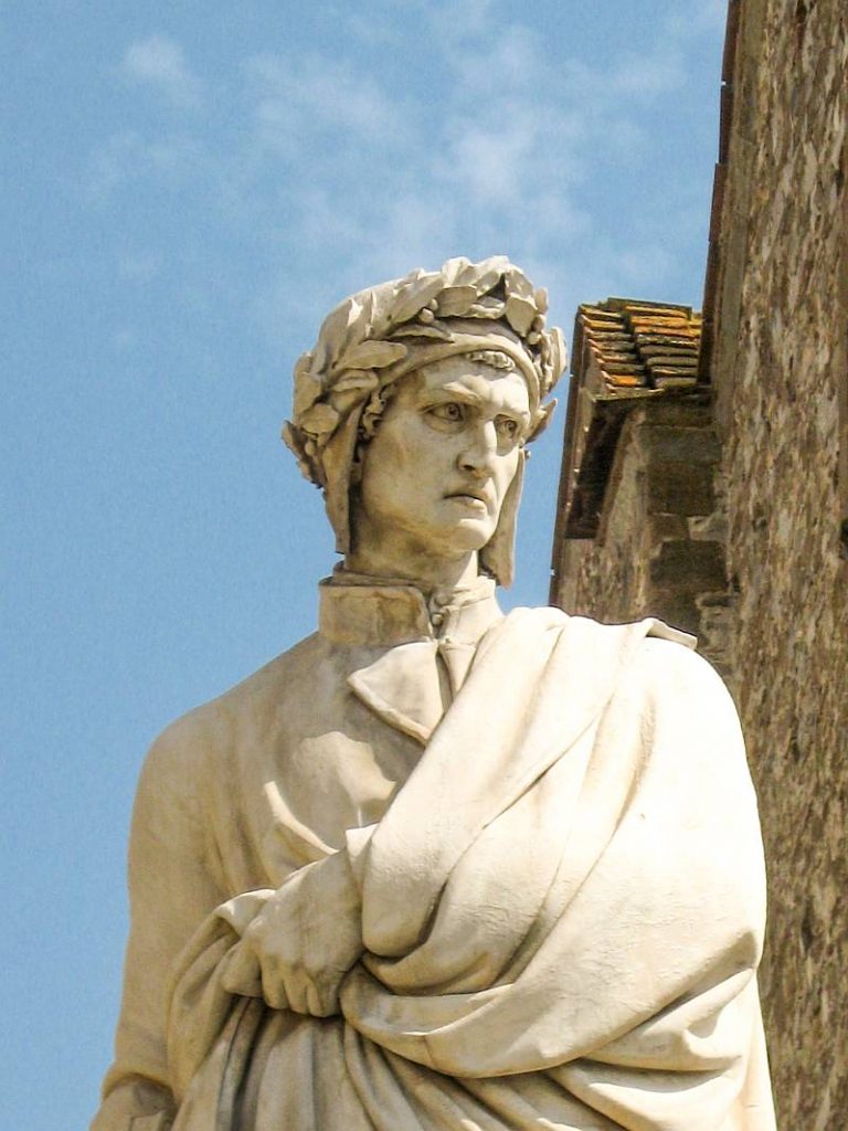 Marble statue of a man wearing a laurel wreath and draped clothing, standing outdoors in front of a stone building under a blue sky with scattered clouds. Statue of Dante Alighieri in Florence