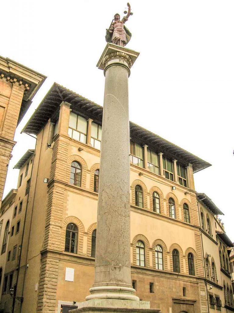 A tall stone column with a statue of a person holding a staff stands in front of a historic building with arched windows and a tiled roof. The Roman Column on Piazza di Santa Trinita