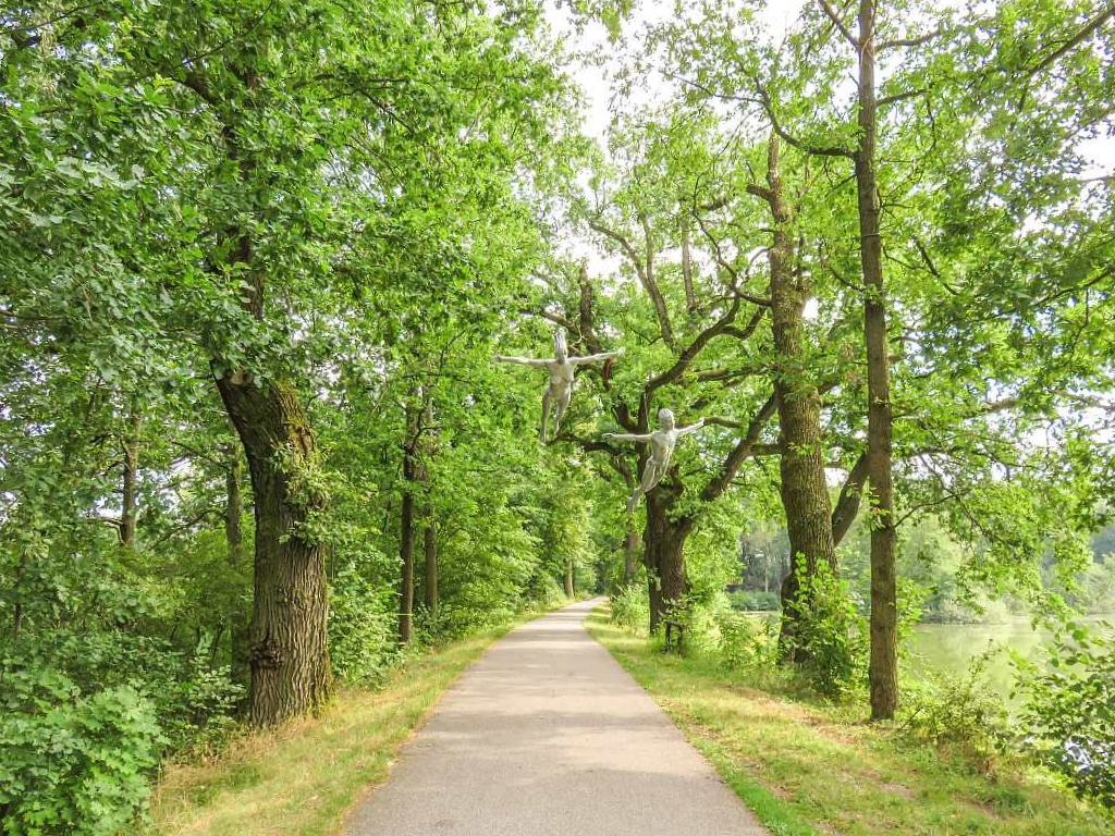 a bike path along a lake lined up with green trees and an art installation of a flying woman and a man; Sport areal Hluboka