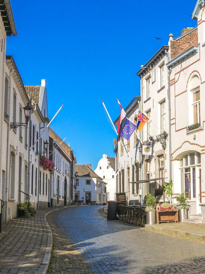 a cobblestone street lined up with white buildings with flag mast on some of them and flags blowing in the wind, on one side some tables and chairs on the sidewalk, part of a cafe, the little white town Thorn in the Netherlands