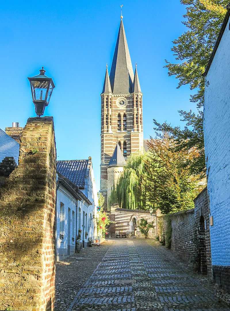 a cobblestone street going up a hill and at the end a church with high belfry, a lot of trees and blue sky
