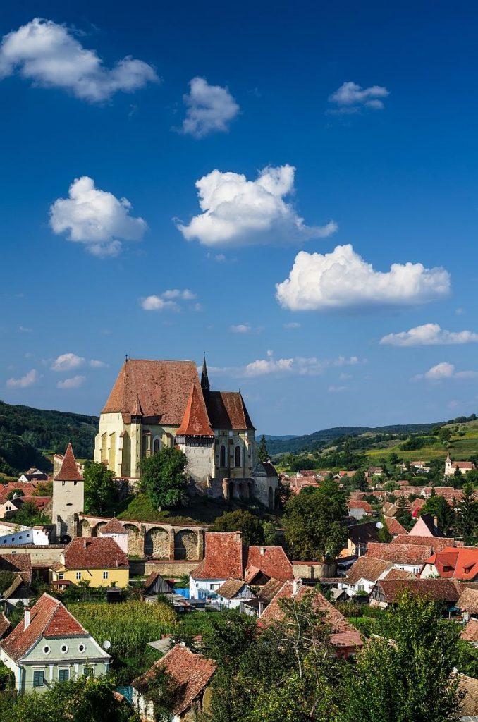 a church on a hill with a village houses around, some green hills at the background and blue sky, Biertan village in Romania