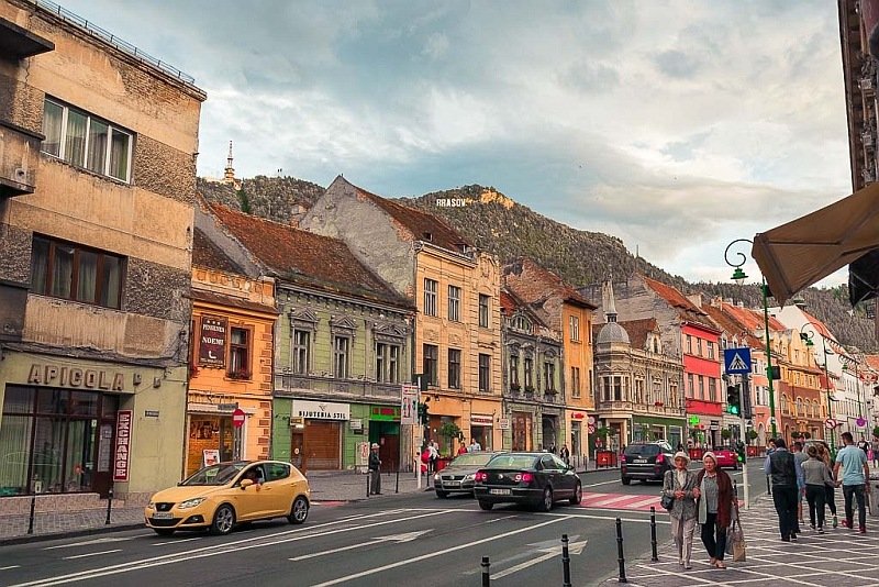 colorful houses in a dilapidated status, people walking in a sidewalk and cars on the street, a hill behind the houses with the letters Brasov on it, Brasov in Romania