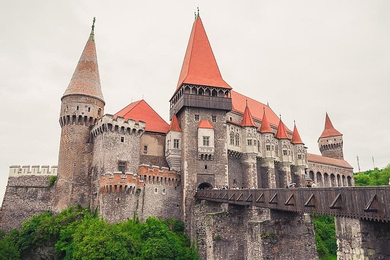 a medieval castle with lots of red-roffed turrets and a long wooden bridge above a gorge leading to the castle, Corvin castle in Transylvania, Romania