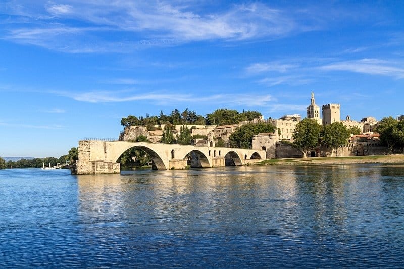 a half bridge over a river with 4 arches and a church tower and a square tower at the background, the bridge of Saint Benezet in Avignon