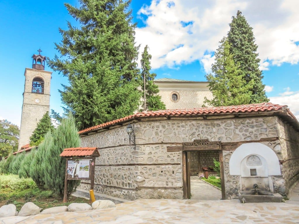 a church with a high stone wall and a gate and clock tower to the right; the Holy Trinity Church in Bansko