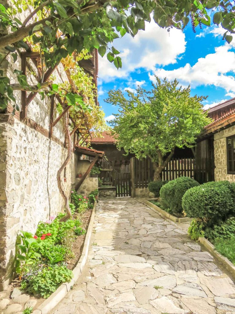a small paved path alongside a house with vine hanging on a wooden pergola; an inner yard of a house in Bansko