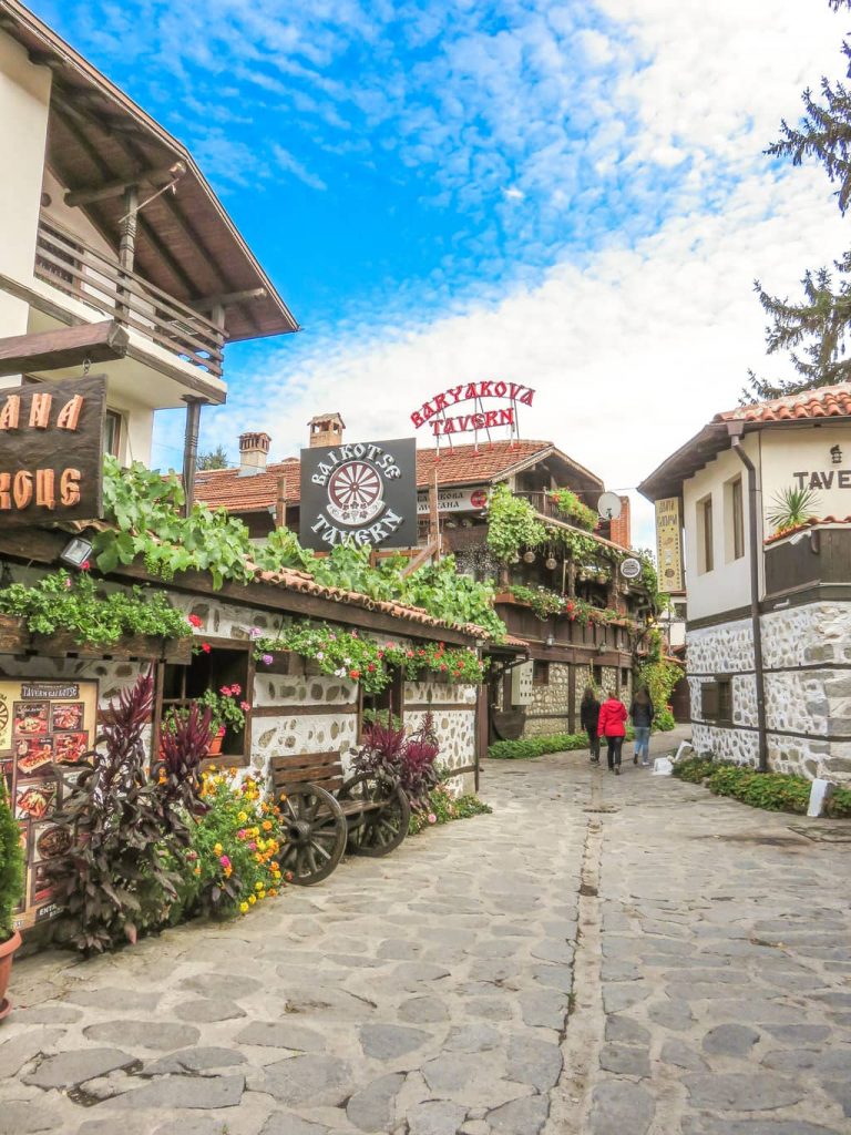 a street with old houses and lots of flowers and signs of local taverns in bansko