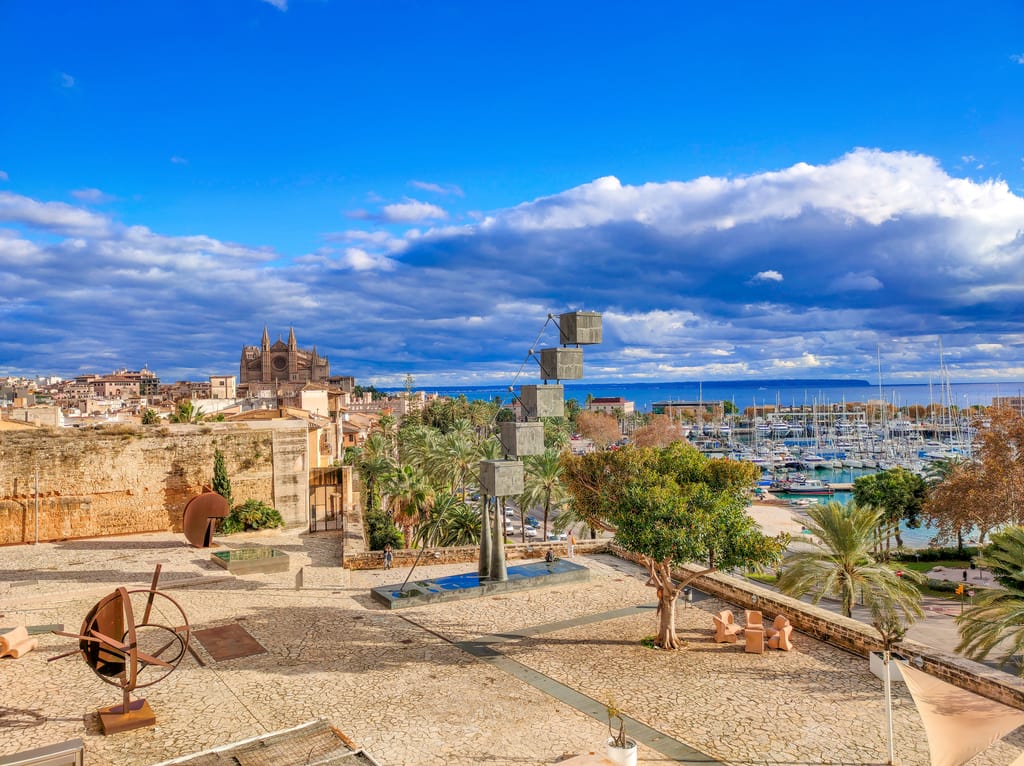a courtyard of a citadel with some modern sculptures and a view to a cathedral and a marina for yachts; view from the Es Baluard museum in Palma de Mallorca