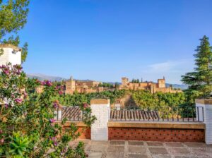 a view from a terrace to a reddish fortress on hill with cypress trees; the Alhambra in Granda