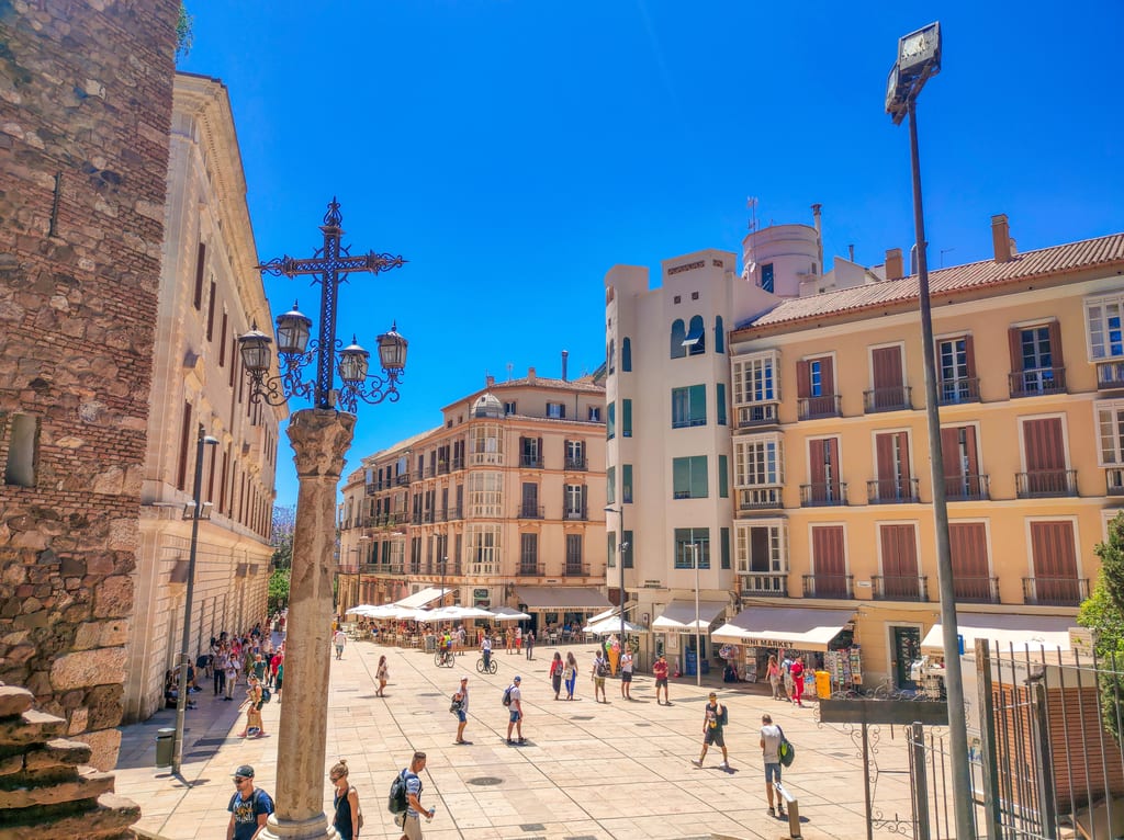a pedestrian street in a city with a column with a cross and four lamps in the foreground; Plaza de la Aguana in Malaga