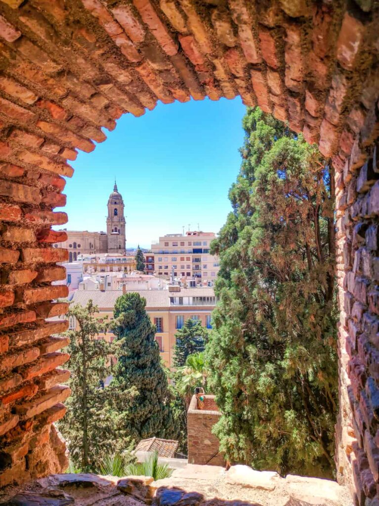 a view from a rounded opening in a fortress towards a cityscape with a cathedral and some green trees; view from the Alcazaba of Malaga fortress