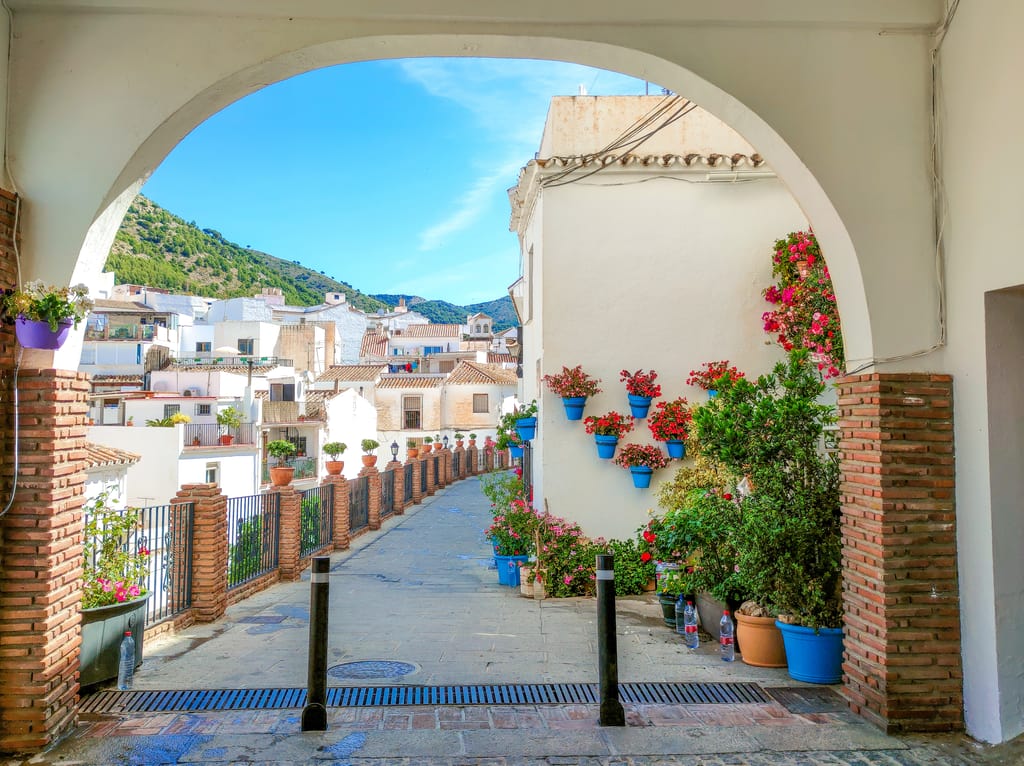 an arch opening a view to a narrow street with white houses on the left side and lots of flower pots. On the right a white houses perched on a hill; a view from Mijas in Spain