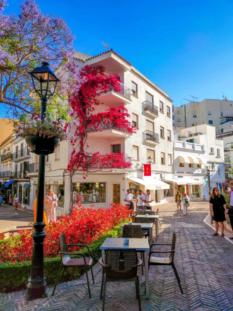 a small square with a few restaurant tables, a lamp pole with flowers hanging on, an a small patch with red flowers; at the background white buildings with climbing plants blooming in red on the facade; Marbella, Andalucia, Spain