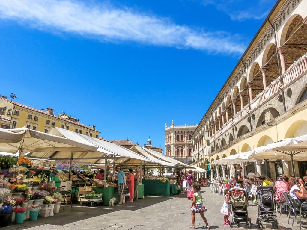 a square with a farmers market and a porticoed building to the right with cafes and people sitting on tables and doing groceries at the market stalls; Piazza delle Erbe Farmer market in Padua