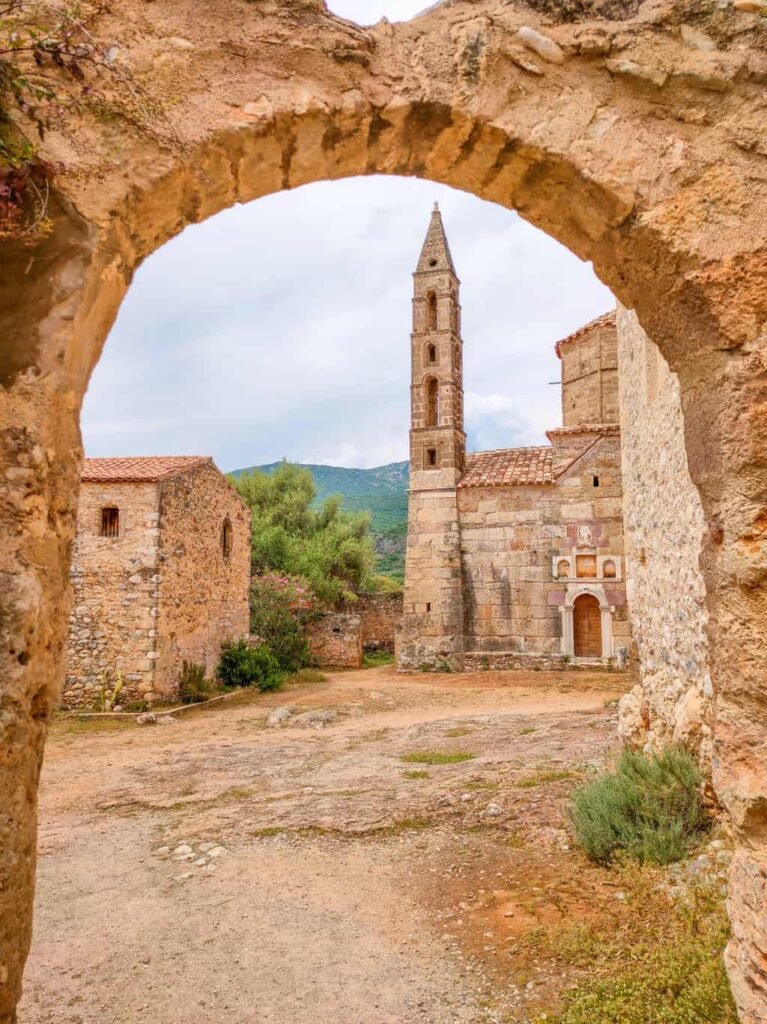An arched stone gateway leading to a small square with a square church tower and some green mountains at the background. The Church of Agios Spyridon in Kardamyli