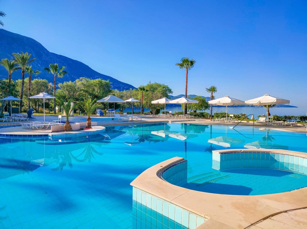 A swimming pool of a resort with palm trees, chaise-lounges, umbrellas and mountain views at the background. Grecotel Filoxenia in Kalamata, Greece