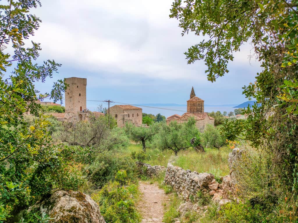 A view with a tower and some stone buildings framed by green with a path leading to the fortress, At the background a see that's blending with the mountains on the horizon. Upper Kardamyli, Old Town Kardamyli in Greece