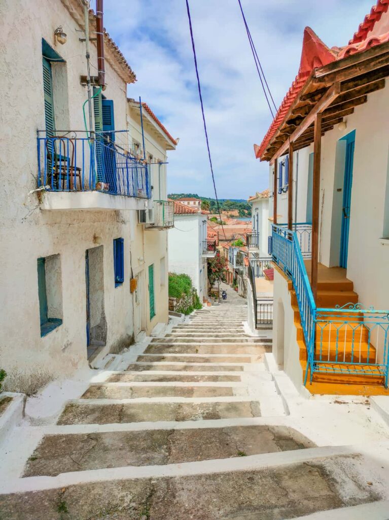 Stone staircase with painted white steps leading down a hill, lined up with white houses with blue or green windows and doors. An alley in Koroni, Greece