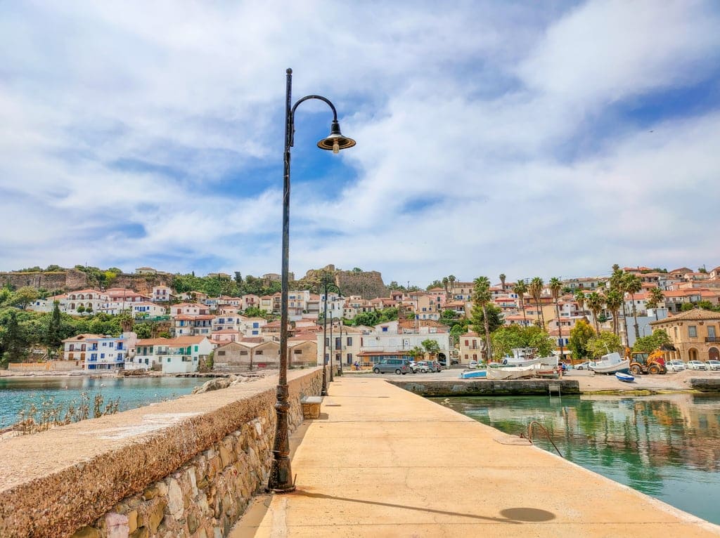 A pier leading to a village built on a low hill with castle ruins on the top of the hill. A view from the pier in Koroni, Greece