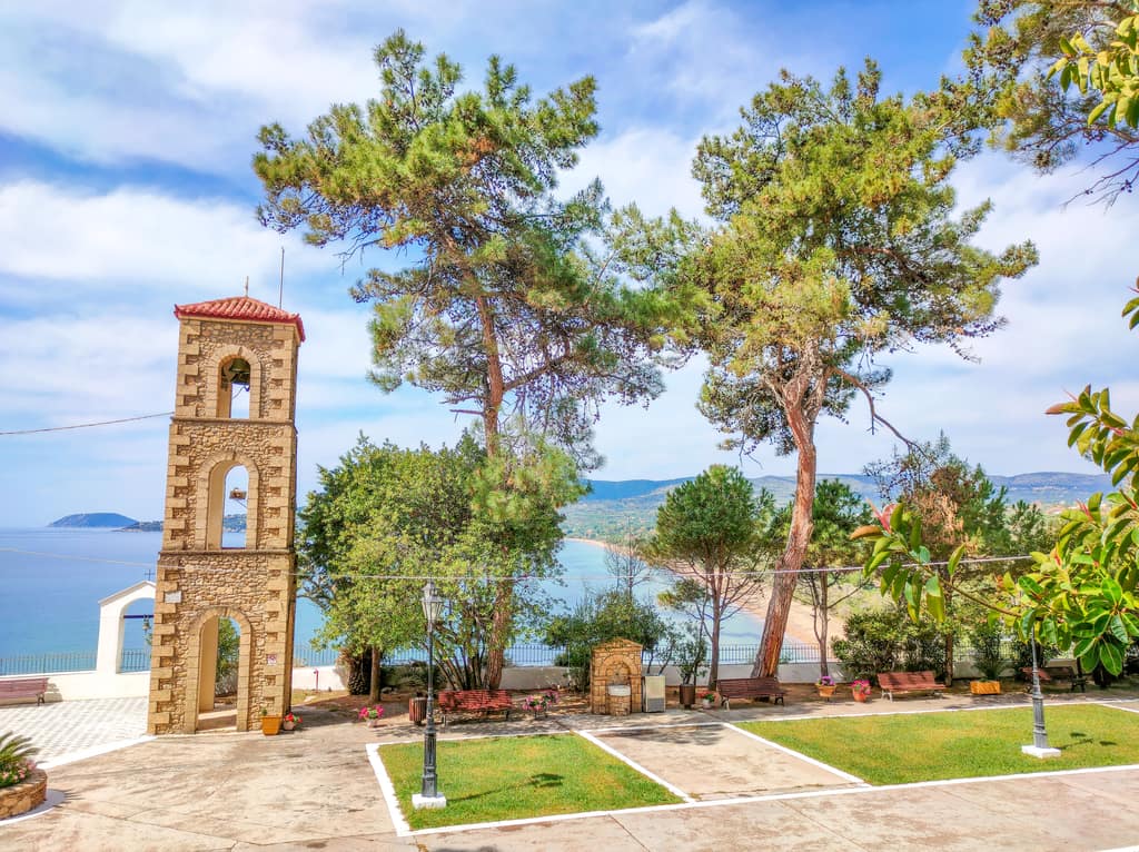 A tree-storey church tower with church bells on top with 2 tall pine trees next to it and some other smaller trees. At the background a bay with a beach and a mountain. The view from the Church of Panagia Elestria in Koroni, Peloponnese, Greece