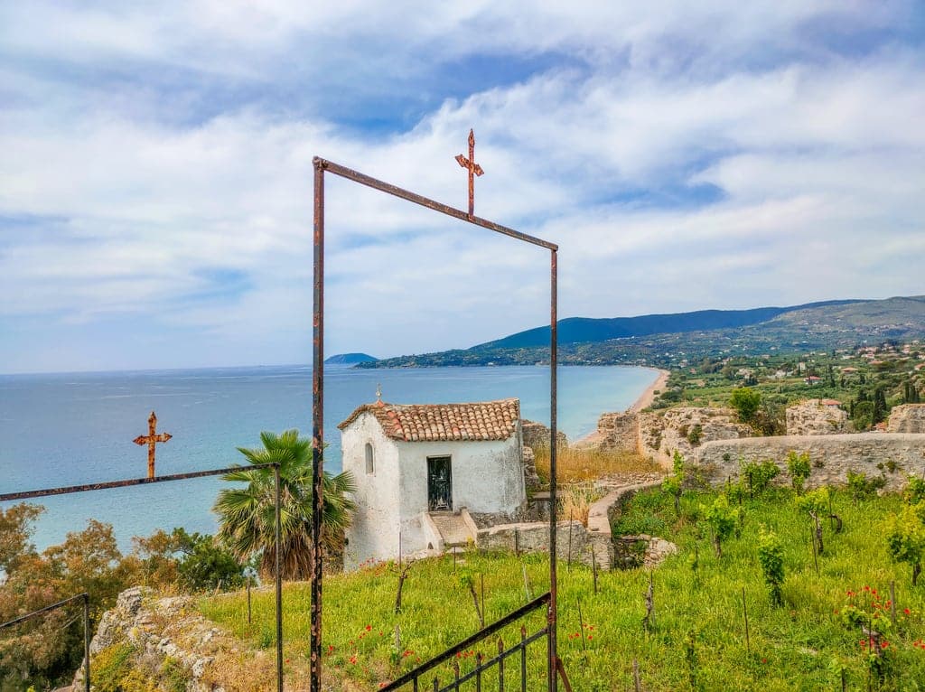 A view from the top of a hill of a bay with some mountains at the background. At the foreground a small white chapel an two metal gates with crosses. The ramparts of a castle are to be seen as well. A view from the Castle of Koroni, Greece