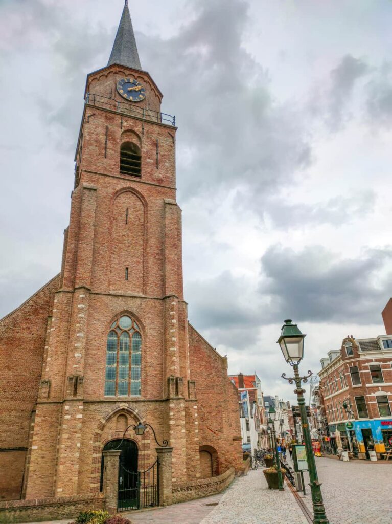 a tall church with a blue clock and a street to the right fo the church; Oude Kerk in Scheveningen