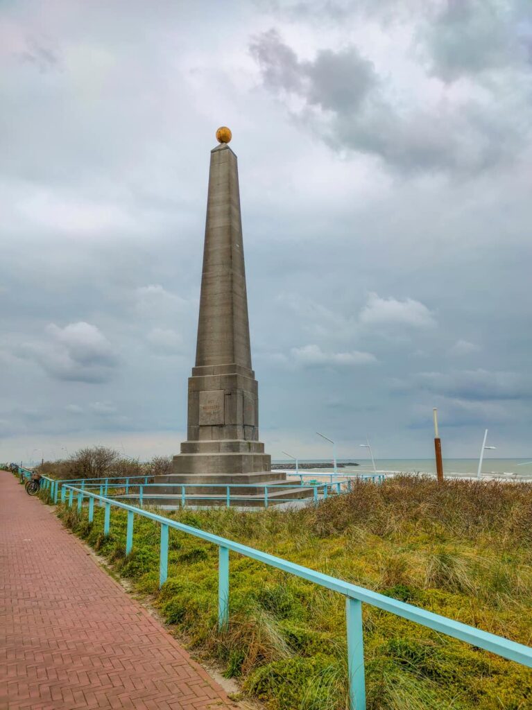 an obelisk with a golden globe on the top, at the background - sea; on a gloomy day with dark clouds; The Memorial Needle in Scheveningen