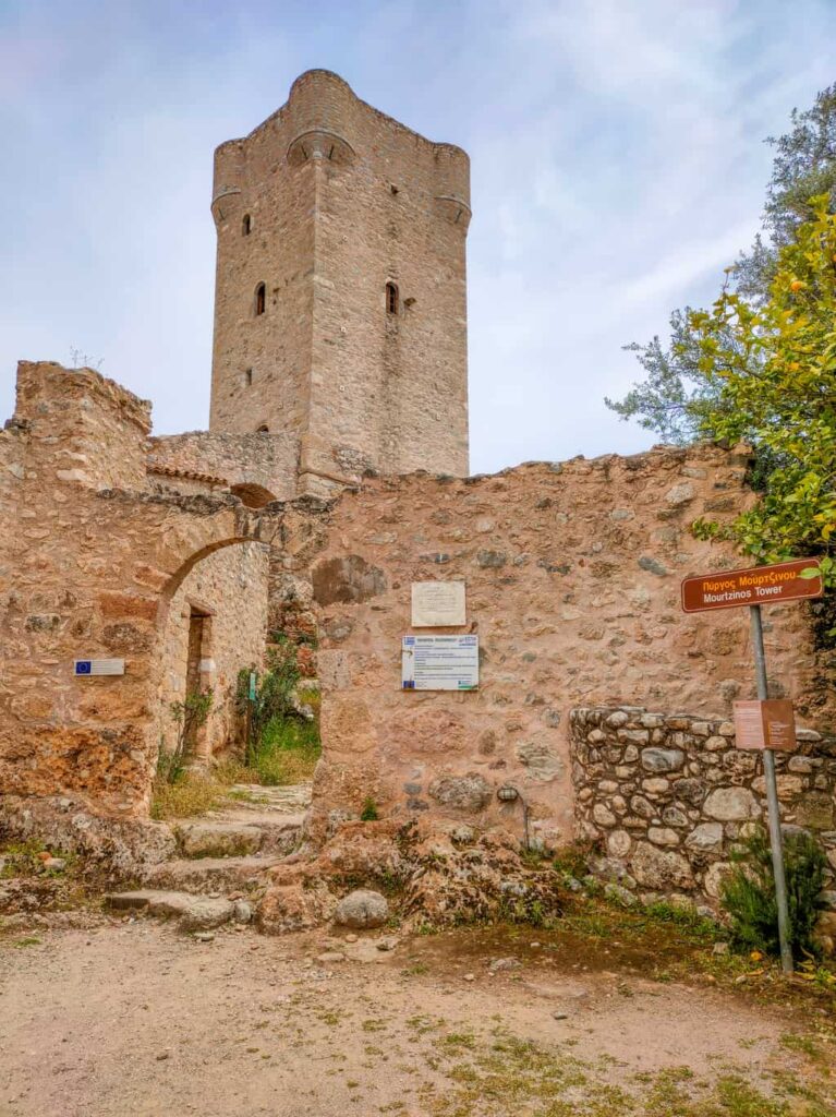 A square tall tower house at the background with a stone wall in front and an arched gateway. THe Mourtzinos Tower in Upper Kardamyli on Peloponnese, Greece