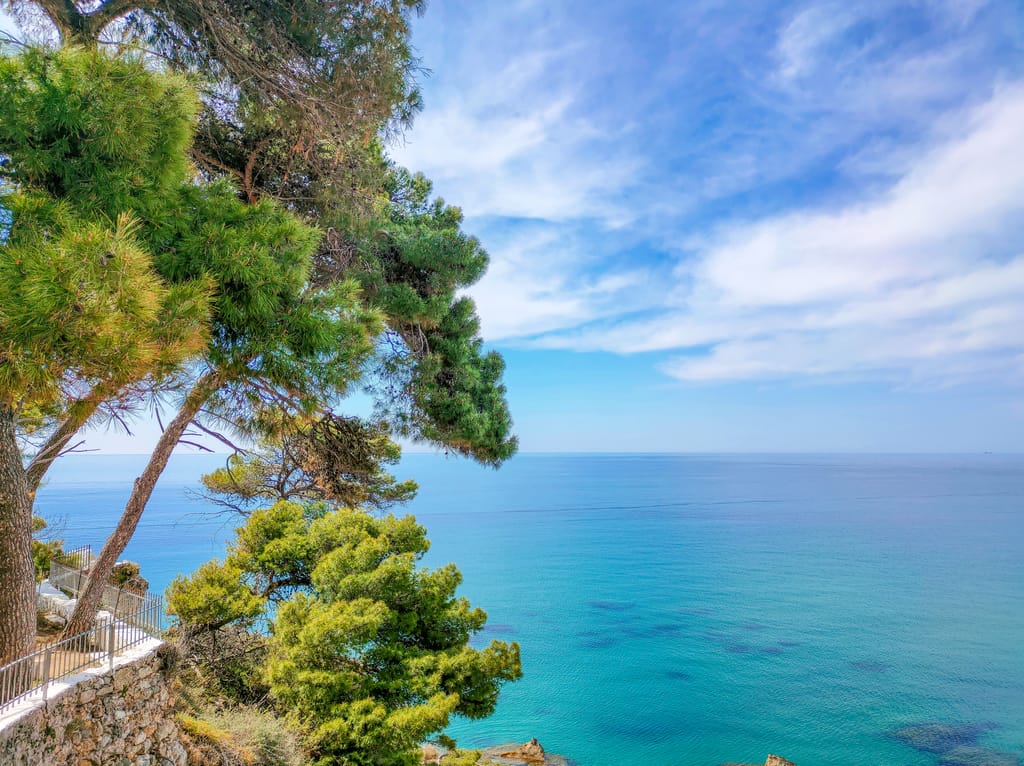 Pine trees framing the blue waters of a sea with blue sky and white clouds. A view of the Messenian Gulf from Koroni, the perfect day trip from Kalamata