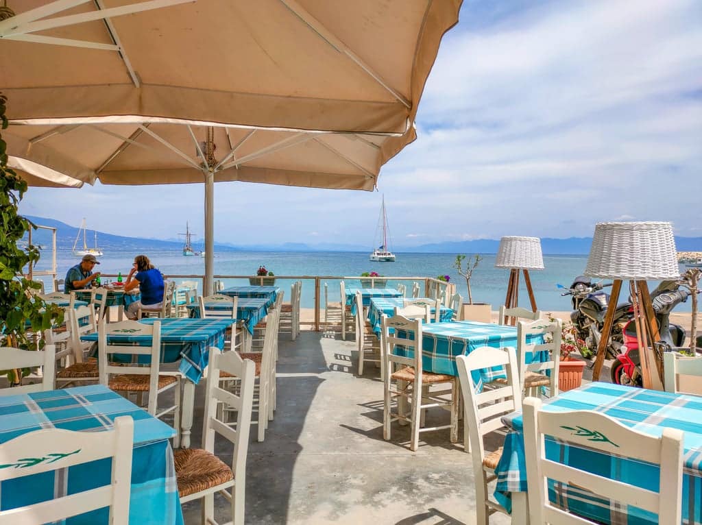 A seaside restaurant featuring an outdoor dining area shaded by large beige umbrellas. Tables are set with blue and white checkered tablecloths. Two people are seated at one of the tables on the left. In the background, you can see the calm sea with a few anchored boats, framed by mountains and a partly cloudy sky. A restaurant in Koroni, Peloponnese