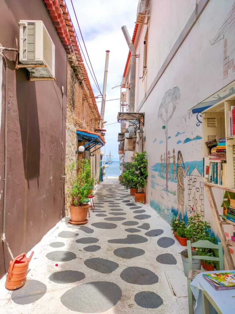 A narrow alleyway with stone pavement, leading to the sea. There are buildings on both sides, with potted plants, air conditioning units, and a colorful mural on the right. A bookshelf with books is visible, and the sea with boats can be seen in the distance. A street in Koroni, Peloponnese