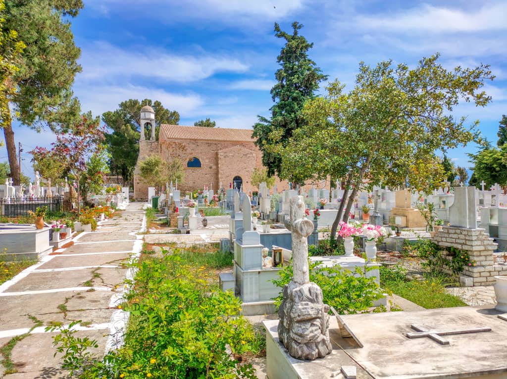 A cemetery with graves, tombstones, flowers, and a church in the background. The Agios Charalambos Church in Koroni Castle grounds