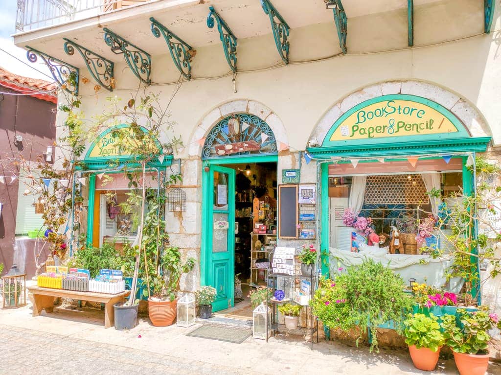 A quaint bookstore with green and turquoise accents, large display windows, potted plants in front on the sidewalk, and a sign reading "Bookstore Paper & Pencil." A bookstore in Koroni, Peloponnese