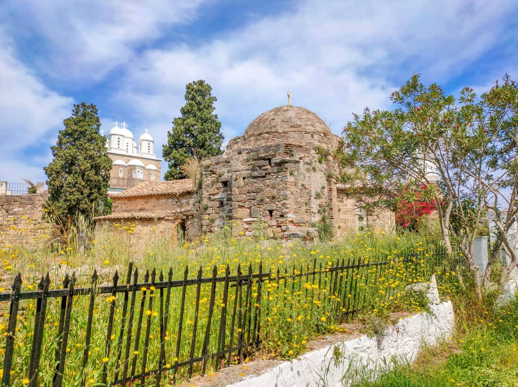 An old stone building with a dome, surrounded by a garden with yellow flowers and a metal fence. In the background, there is a white church with multiple domes and crosses, partially obscured by trees. The sky is clear. Hagia Sophia and Timos Podromos, Koroni