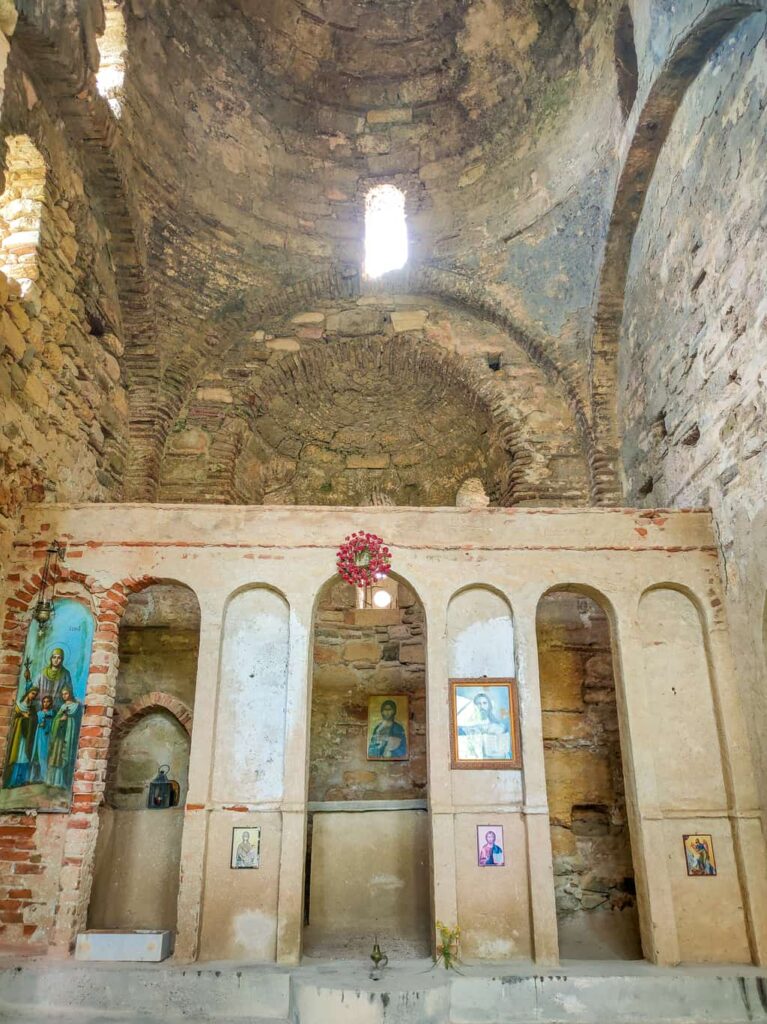 The interior of an old stone chapel with an arched ceiling and weathered walls. the iconostas display religious icons and paintings of saints, and a wreath hangs above the central arch.
