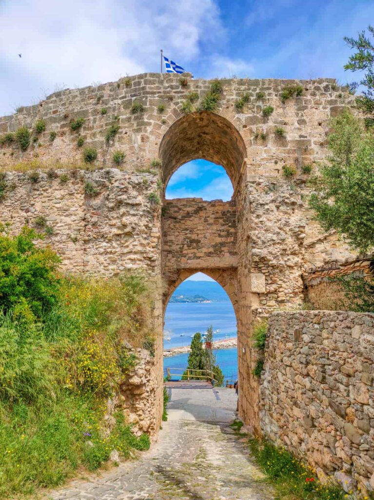 A historic stone archway with the Greek flag at the top, offering a view of the sea through the cobblestone path. The Castle of Koroni