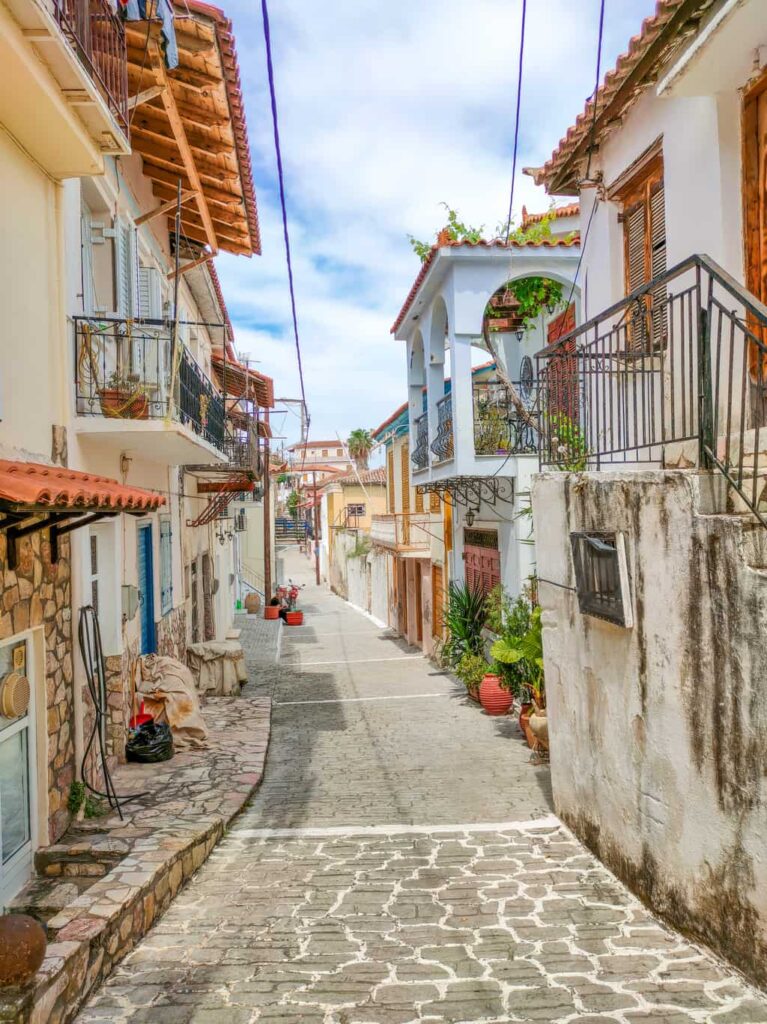 A cobblestone street bordered by Mediterranean-style buildings, with balconies, plants, and clear skies. A tranquil yet lively setting. A street in Koroni