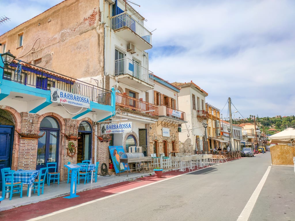 A street in a coastal town lined with Mediterranean-style buildings, restaurants, and parked cars. One restaurant, "BARBAROSSA Fish Restaurant," stands out with its blue signage and outdoor seating featuring blue tables and chairs. The sky is partly cloudy, and hills are visible in the background. A street with restaurant in Koroni
