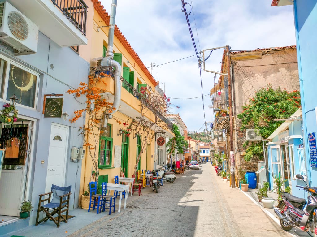 A lively Mediterranean street with pastel-colored buildings, cobblestone pavement, balconies adorned with flowers, and outdoor seating. A street in Koroni