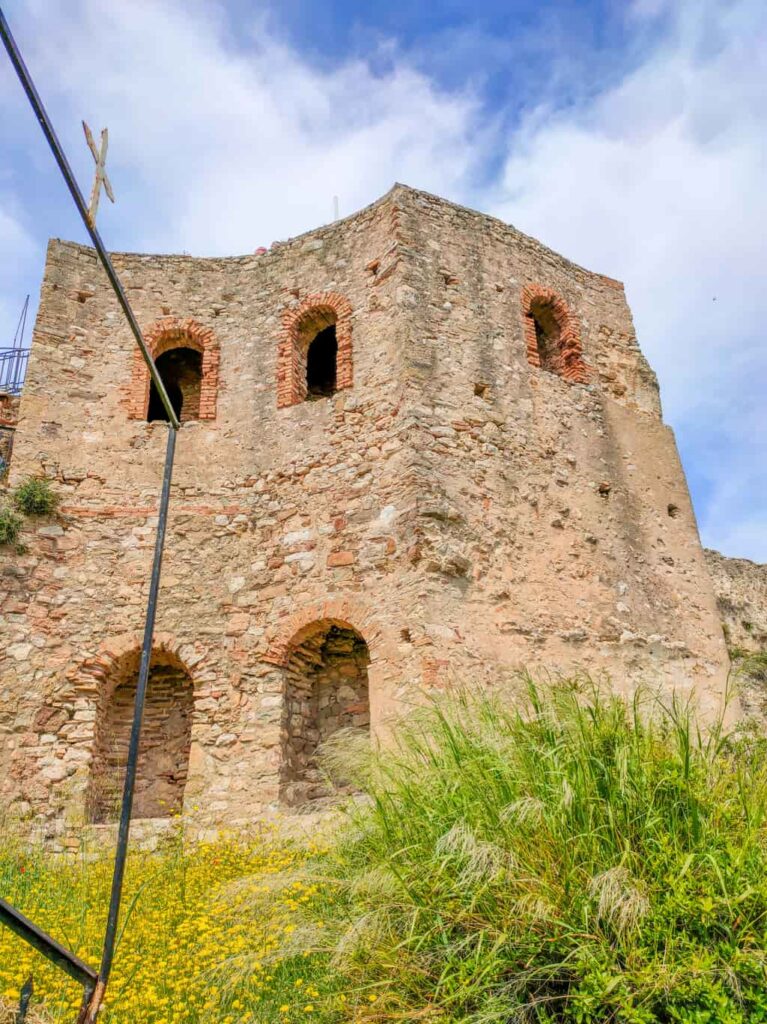 An aged stone building with arched brick-framed openings. A metallic structure with a cross is present nearby. The foreground showcases green grass and yellow flowers, with a partly cloudy sky above. The Octagonal Tower at the fortress in Koroni