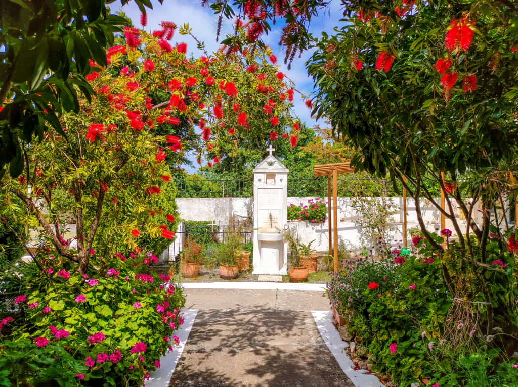 A vibrant garden featuring colorful flowers, green plants, and a pathway leading to a white shrine with a cross. A peaceful and lush setting. The Timios Prodromos Monastery in Koroni